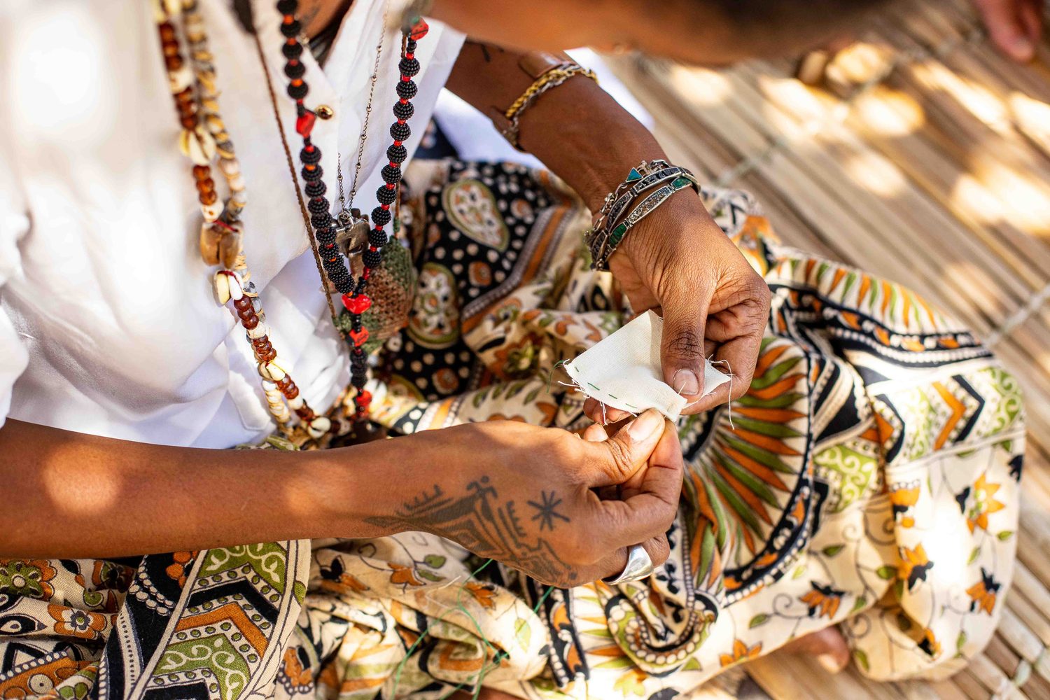Mãos de Yasmim preparando ervas sobre tecido de estampa africana, com guias de contas ao redor